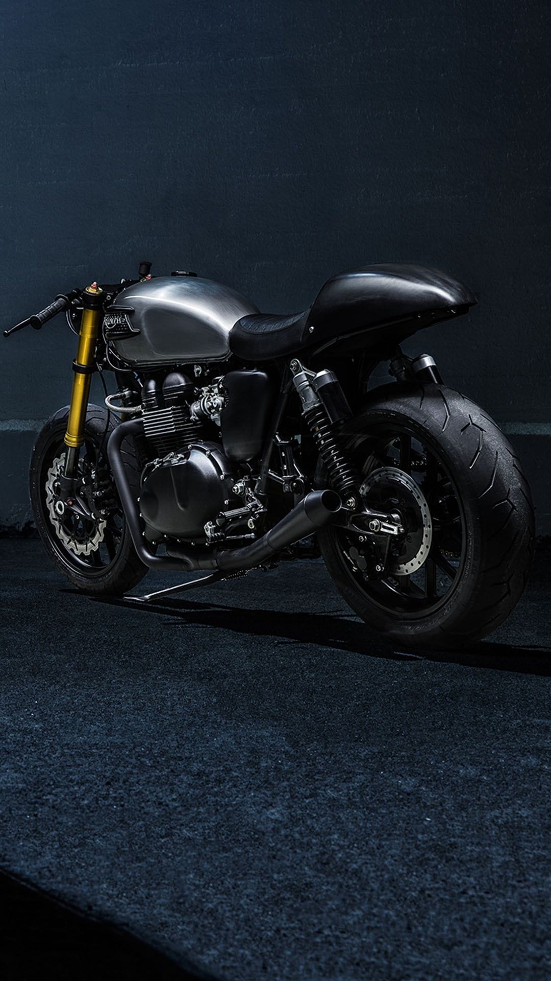 Custom cafe racer motorcycle with a metal fuel tank, gold forks, and black leather seat against a dark wall.