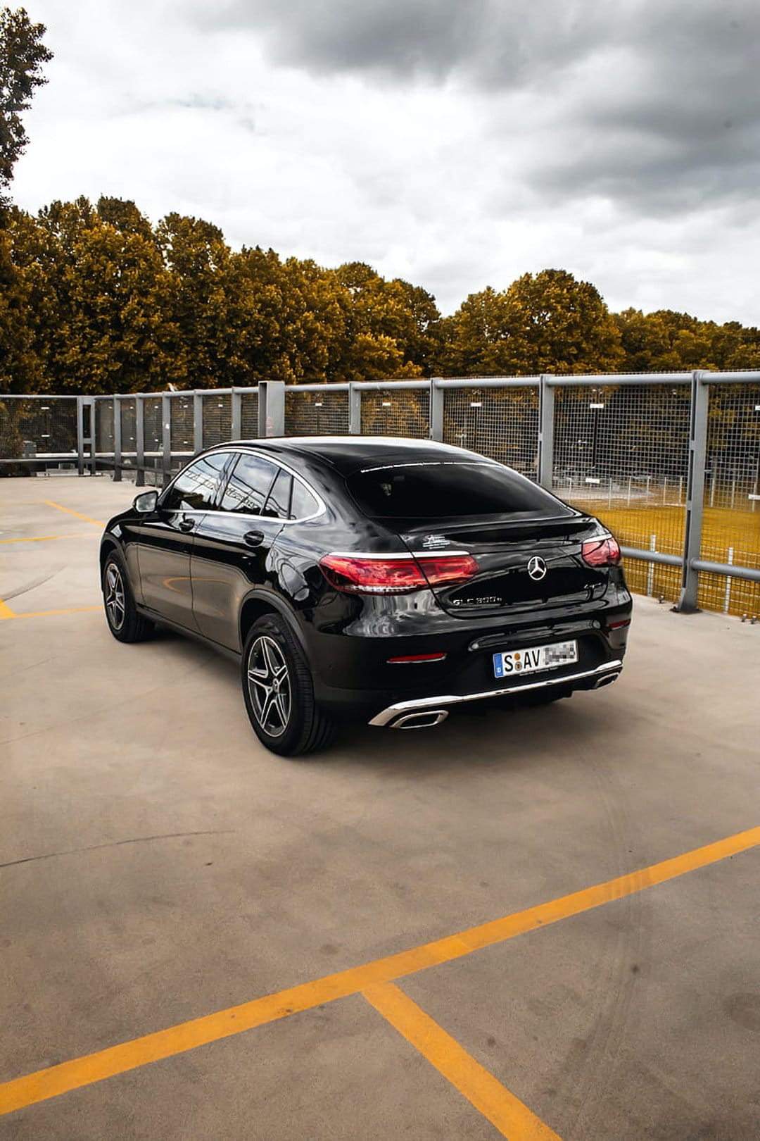 Mercedes GLC Coupe in black parked on concrete with yellow lines, reflecting autumn trees and a cloudy sky.