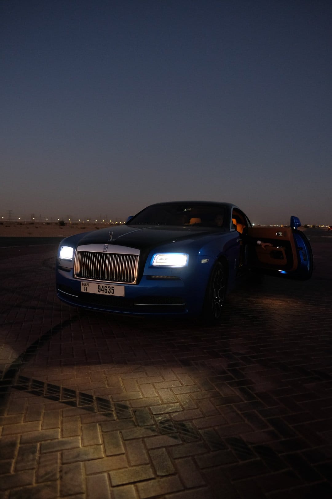 Blue Rolls-Royce coupe with an open door sits on a paved road against a twilight desert and distant city lights.