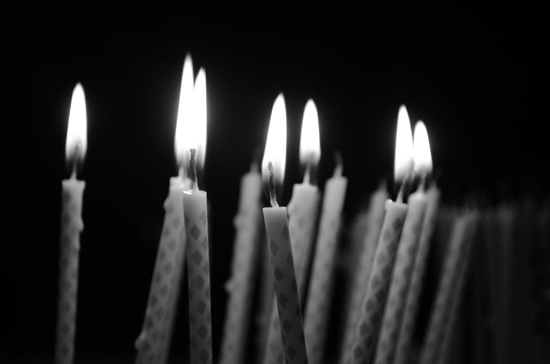 Flickering birthday candles with textured wax patterns glow brightly against a deep black void in monochrome.