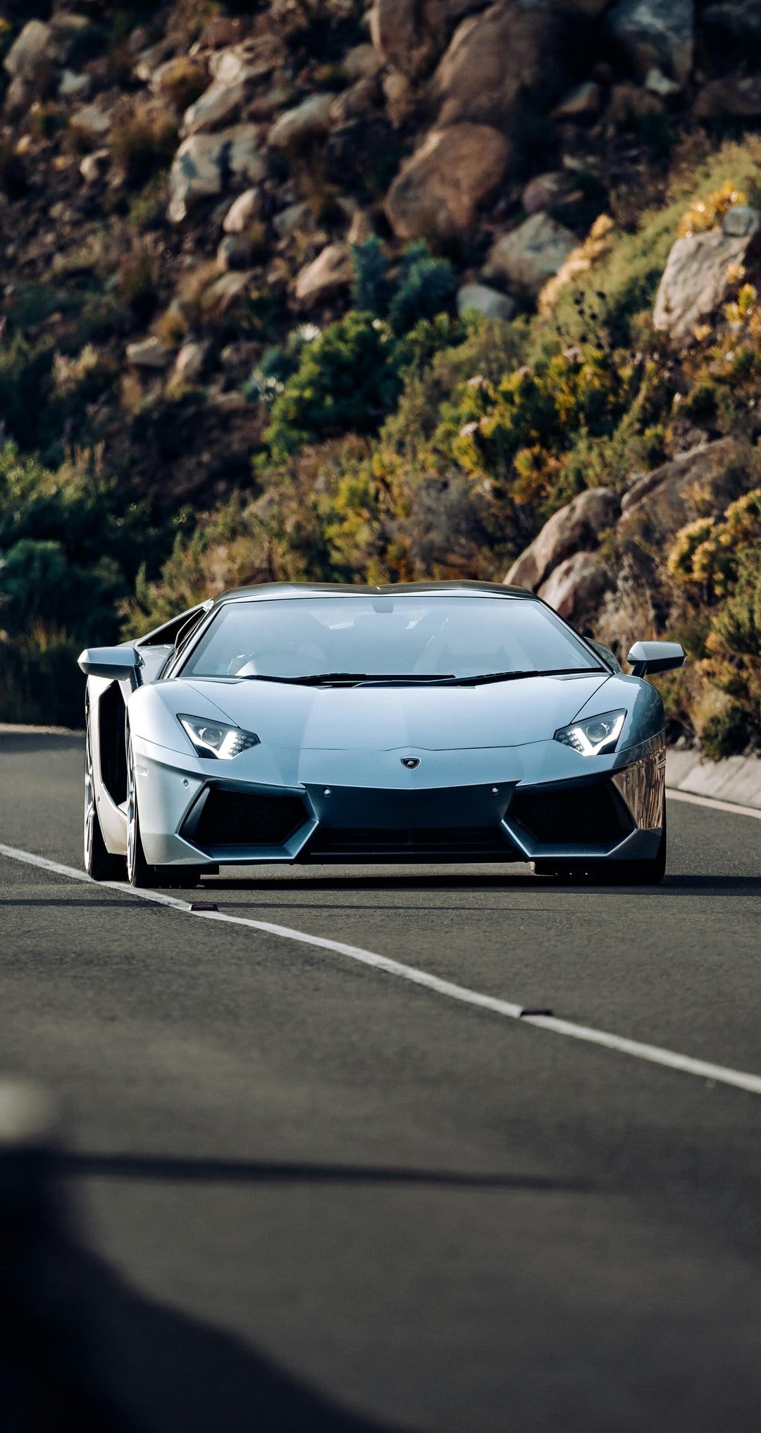 Silver Lamborghini Aventador drives on a winding asphalt road through a rocky hillside under bright sunlight.
