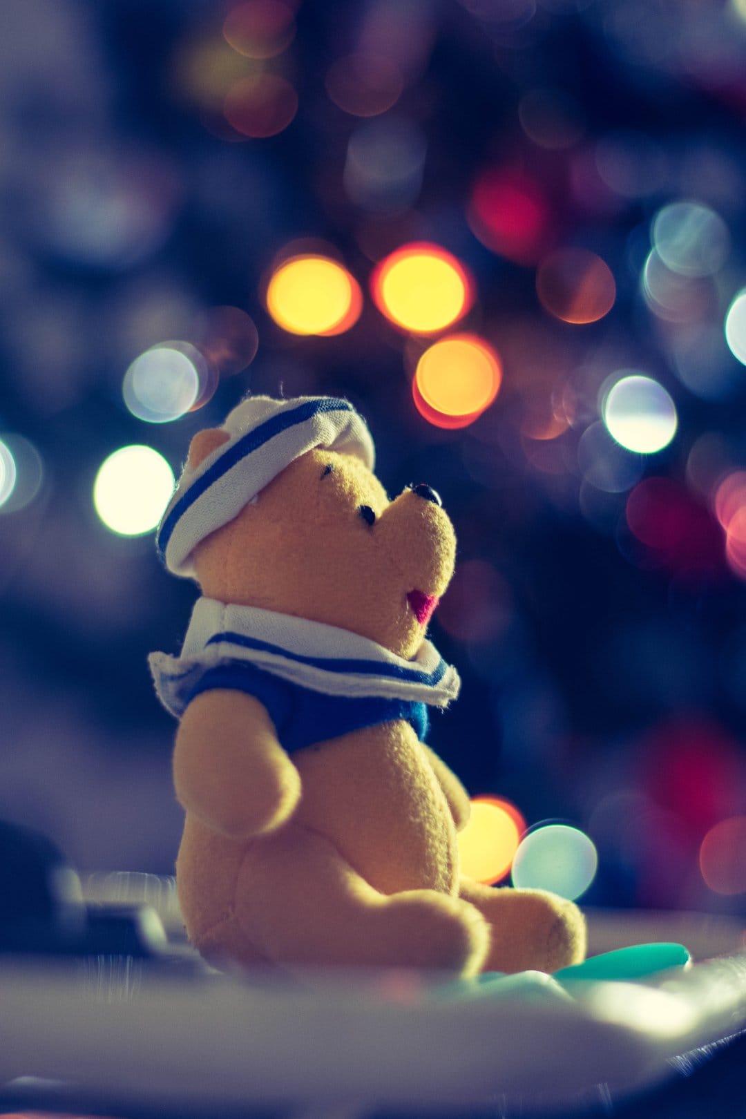 Winnie the Pooh plush in a blue sailor hat and scarf sits before colorful, out-of-focus festive bokeh lights.