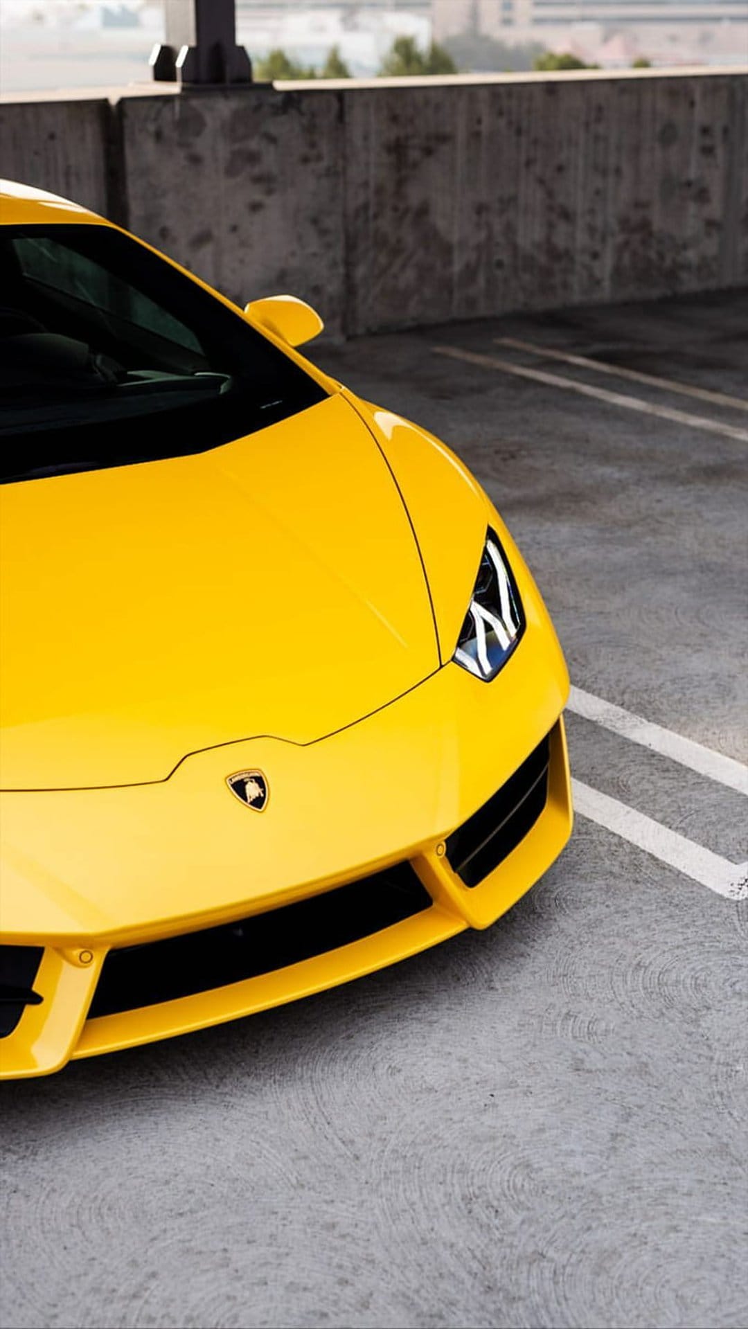 Yellow Lamborghini supercar front view on gray concrete pavement against a tall textured brutalist wall.