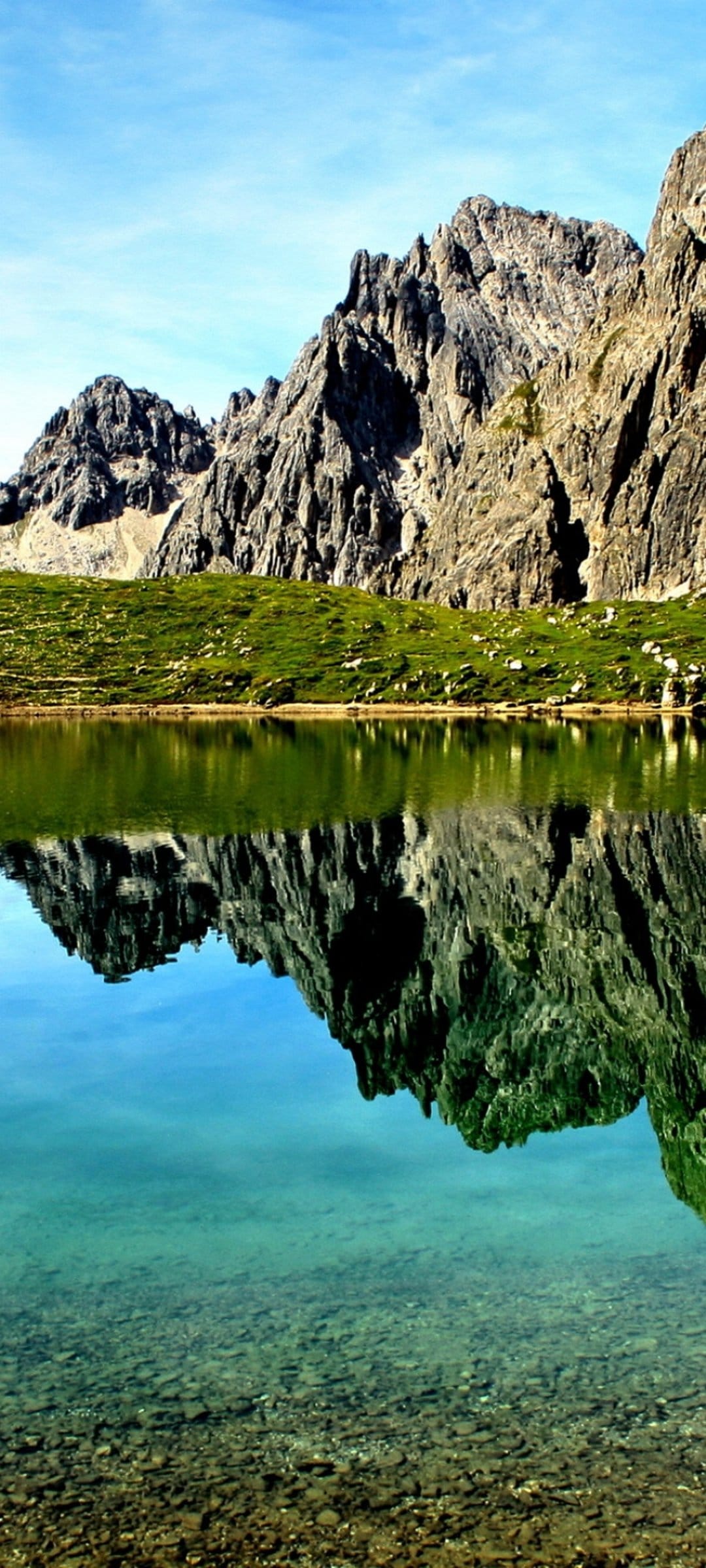 Rugged grey mountain peaks reflect symmetrically in a clear alpine lake under a bright blue summer sky.