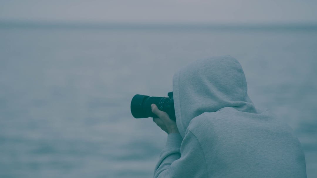 Photographer in a grey hoodie stands at the water's edge holding a camera toward a misty, overcast ocean horizon.