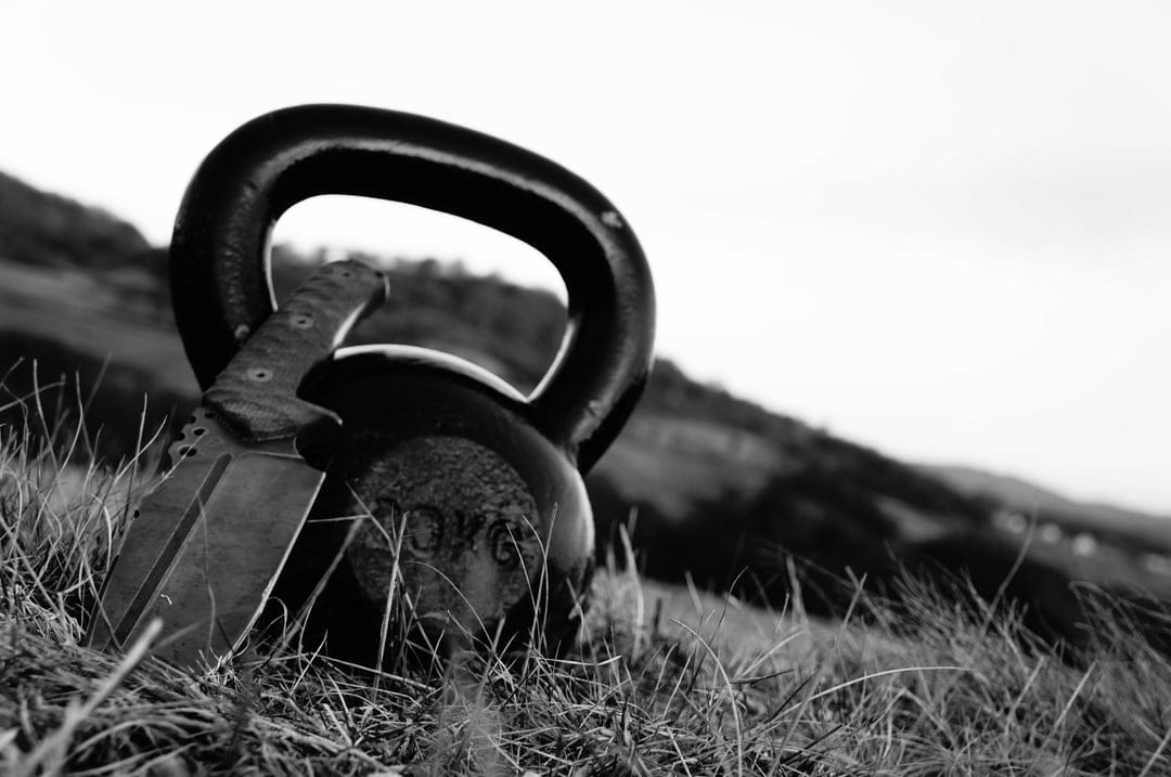 Kettlebell with a knife stuck inside sits in a grassy field with misty hills in a black and white photograph.