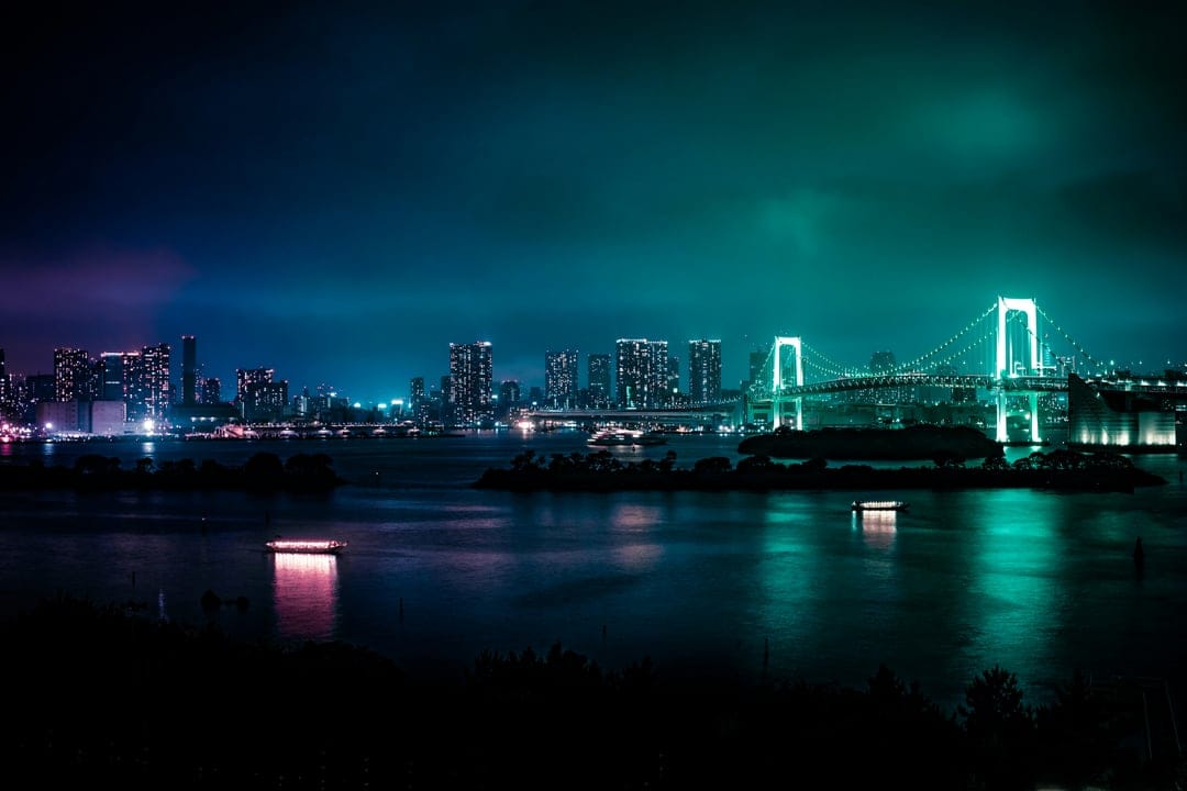 Tokyo Rainbow Bridge glows with turquoise neon lights above dark water reflecting a distant city skyline at night.