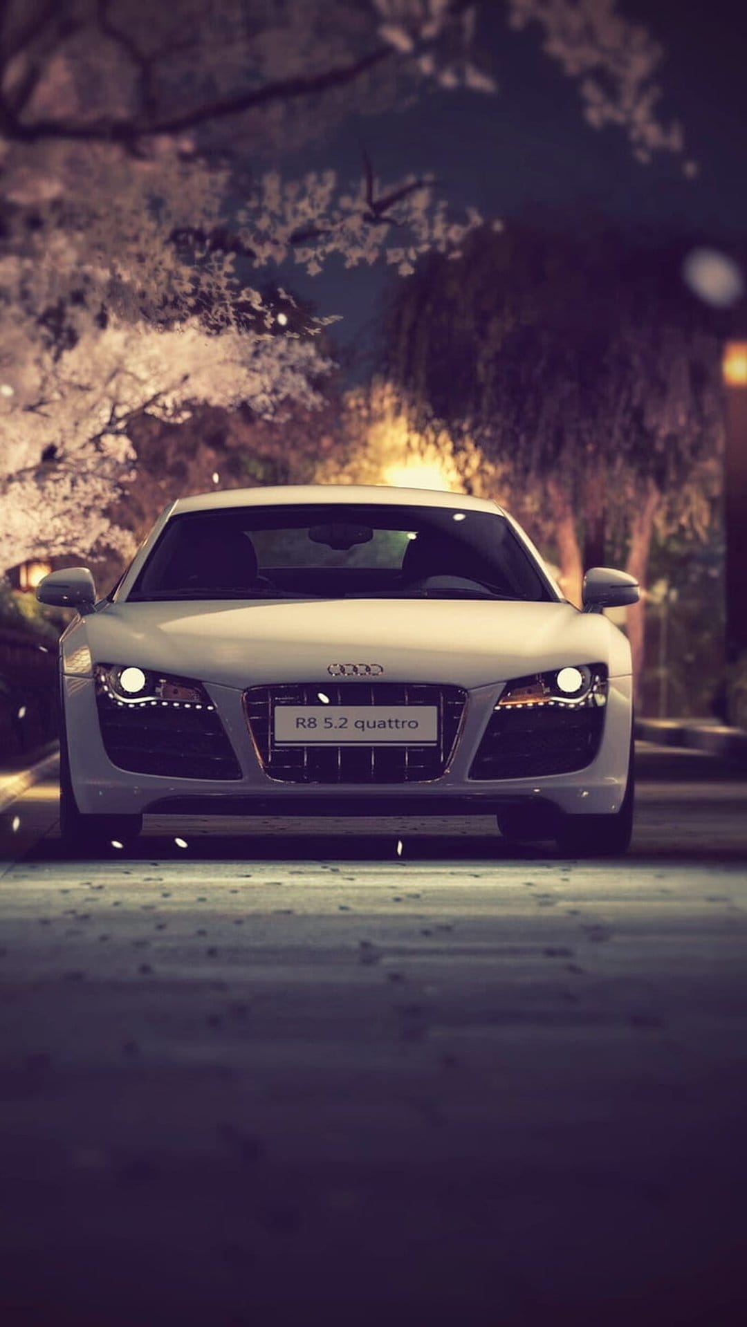 White Audi R8 Quattro parked under white blooming trees at night with glowing headlights and soft street lighting.