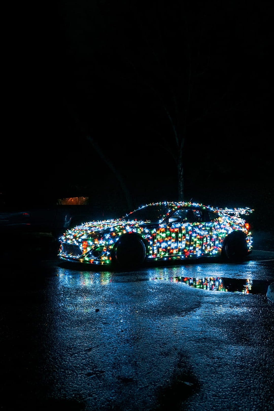 Sports car covered in multicolored glowing Christmas lights reflecting on wet asphalt during a dark night.