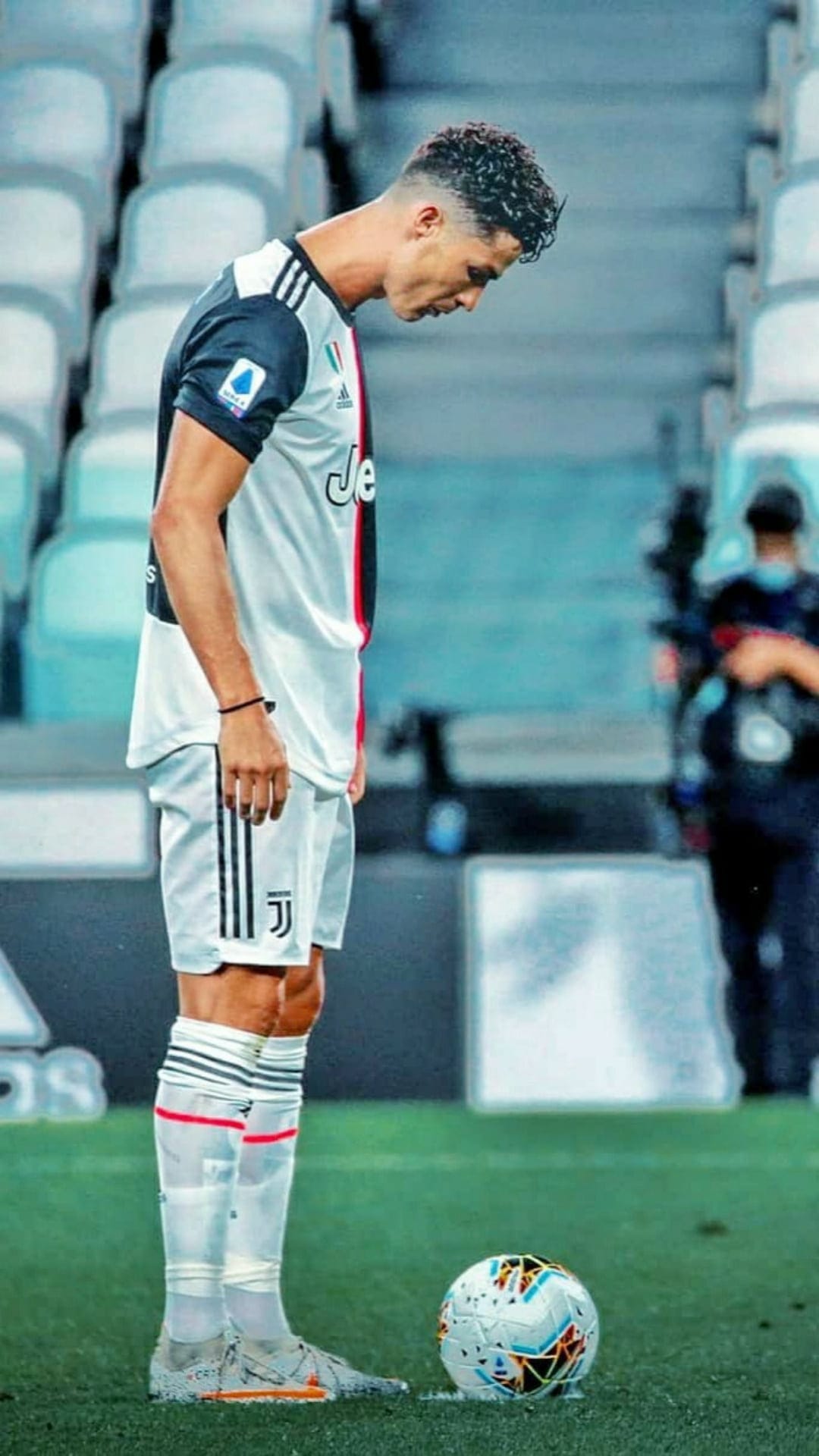Cristiano Ronaldo in a Juventus kit looks at a soccer ball on a green field with stadium seating behind him.
