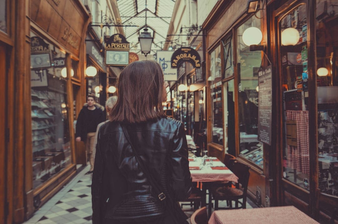 Woman in a black leather jacket walks through a Parisian arcade with checkered floors and ornate street lamps.