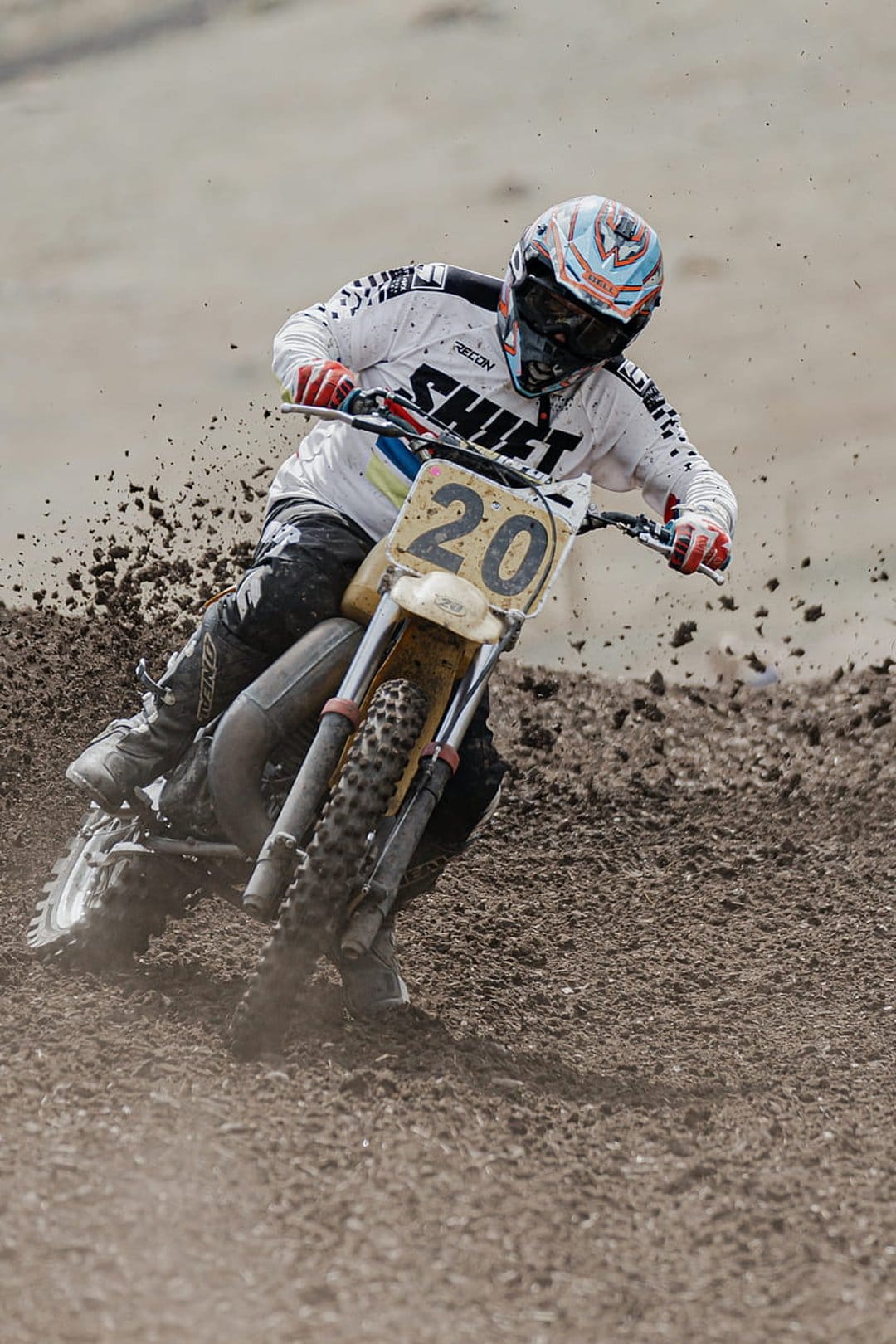 Motocross rider in a white jersey leans into a sharp turn while kicking up dirt on an outdoor racing track.