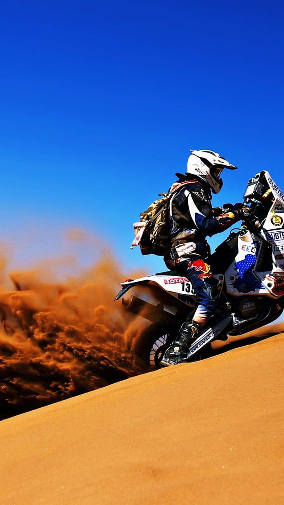 Motorcyclist in racing gear kicks up a spray of orange sand while riding a dirt bike across a desert dune.