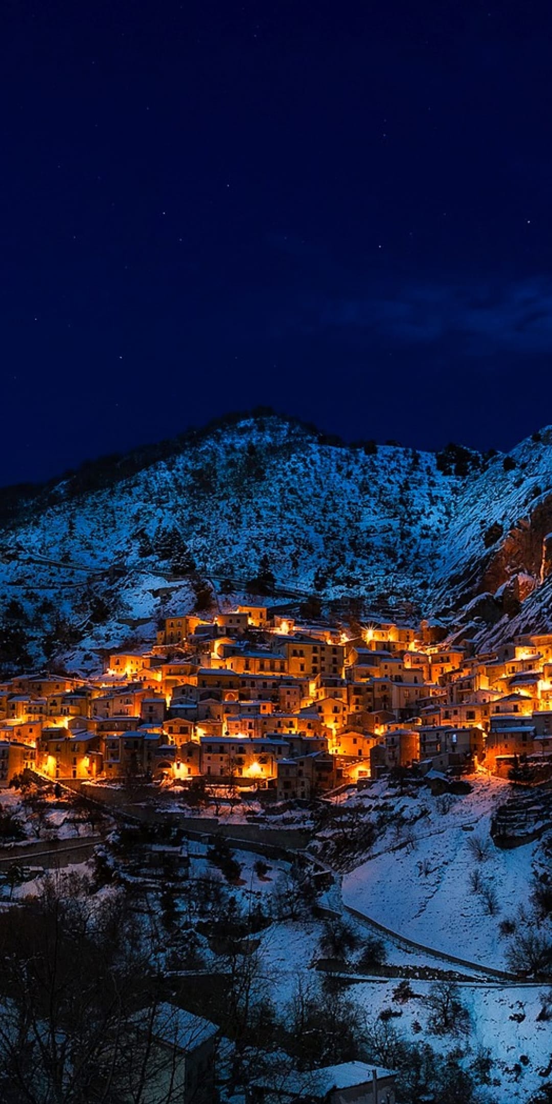 Castelmezzano village lights glow against snow-covered mountains under a dark night sky with visible stars.