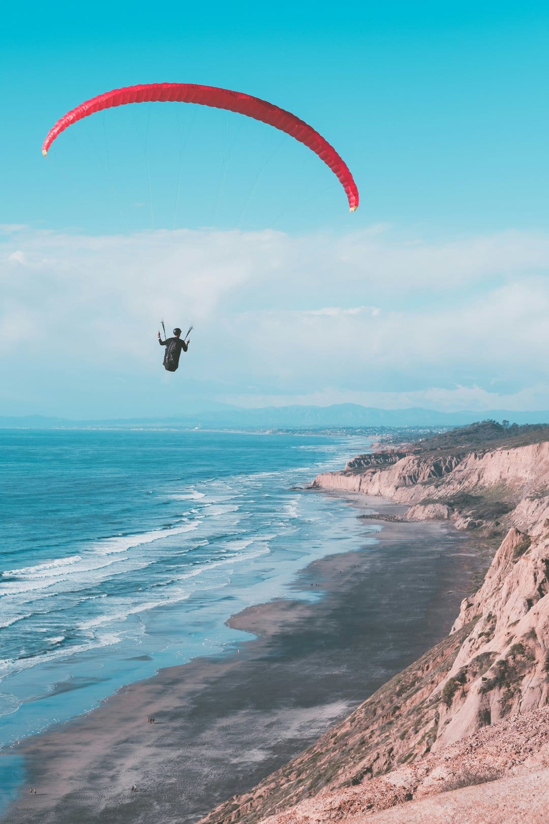 A red paraglider flies high above steep coastal cliffs and turquoise ocean waves under a clear blue sky.