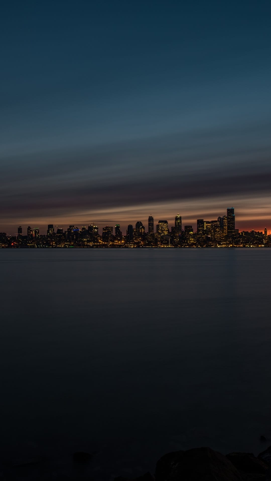 Luminous city skyscrapers overlook a dark harbor under a fading orange horizon during late evening twilight.