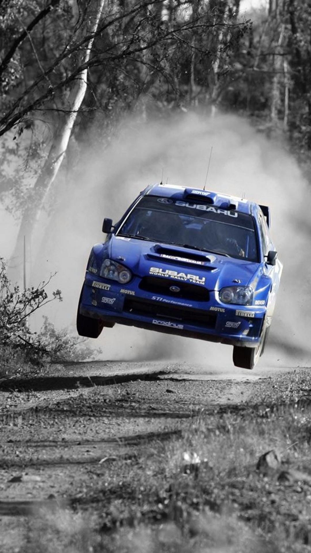 Blue Subaru Impreza WRC rally car jumping over a dirt road with dust clouds and a monochrome forest backdrop.