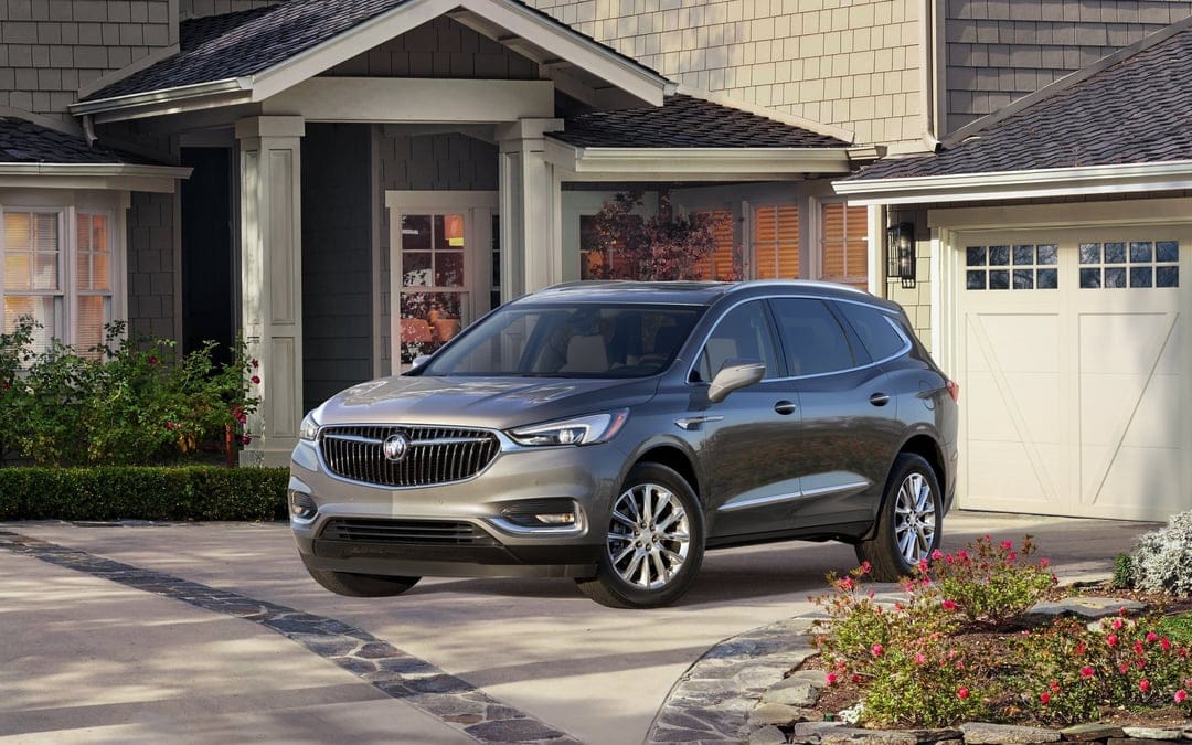Gray Buick Enclave SUV parked in a stone driveway of a modern suburban house with green landscaping.