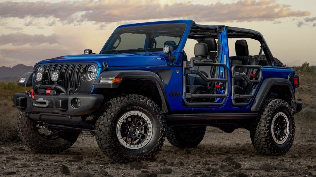 Blue Jeep Wrangler with tube doors and rugged mud tires on a rocky desert floor under a soft sunset sky.