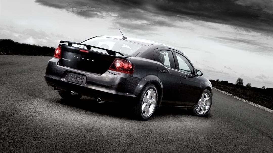 Rear view of a black Dodge Avenger sedan on an asphalt road under a dramatic, cloudy sky with red tail lights.