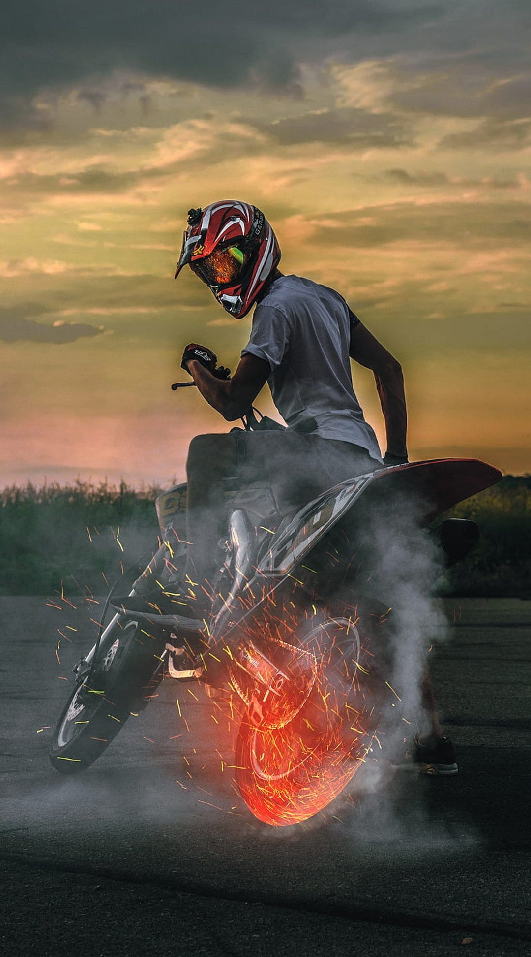 Motorcyclist in a helmet performs a burnout with glowing sparks and thick smoke against an orange sunset sky.