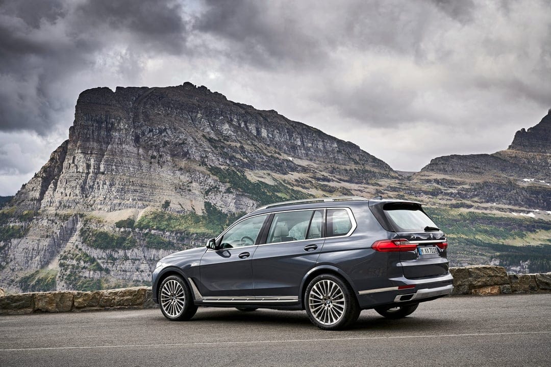 Grey BMW X7 luxury SUV parked on a high mountain road with massive rock cliffs and a cloudy sky background.