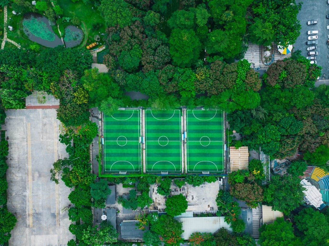 Three green soccer fields with white markings are surrounded by a dense forest from a top-down aerial perspective.