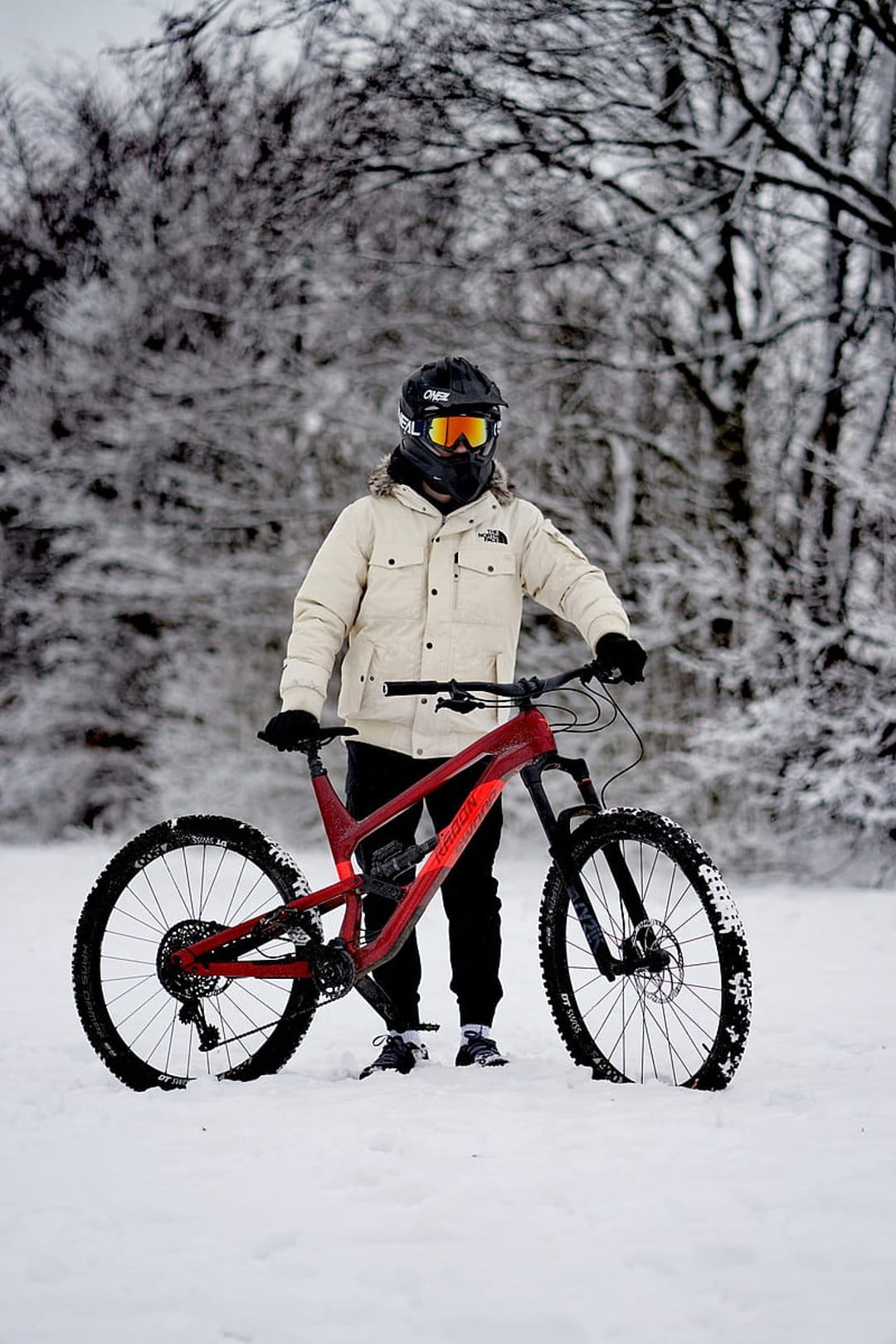 Cyclist in a white puffer jacket stands with a red mountain bike among frost-covered trees in deep snow.