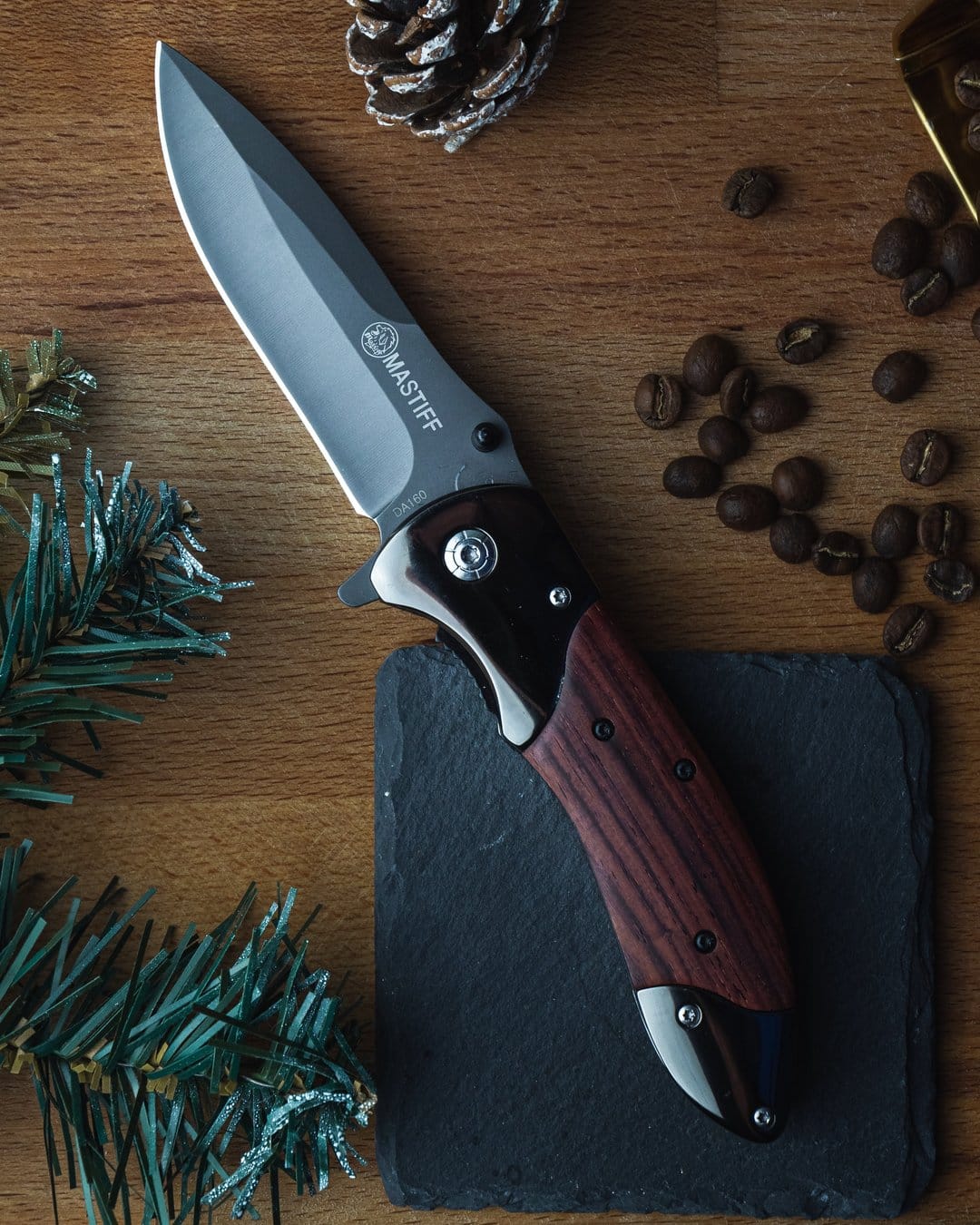 Folding knife with a wooden handle on a dark slate coaster surrounded by coffee beans and fresh pine needles.