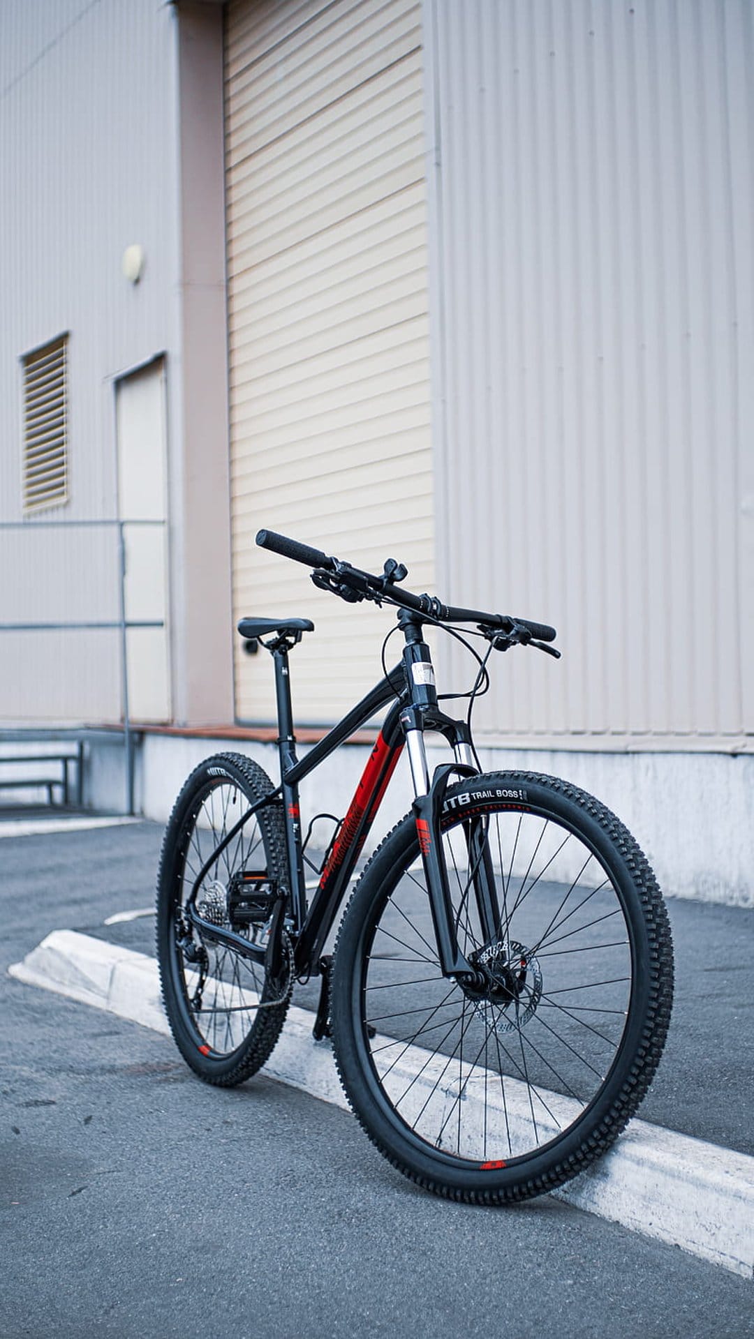 Black mountain bike with red trim and knobby tires standing on asphalt against a corrugated metal wall.