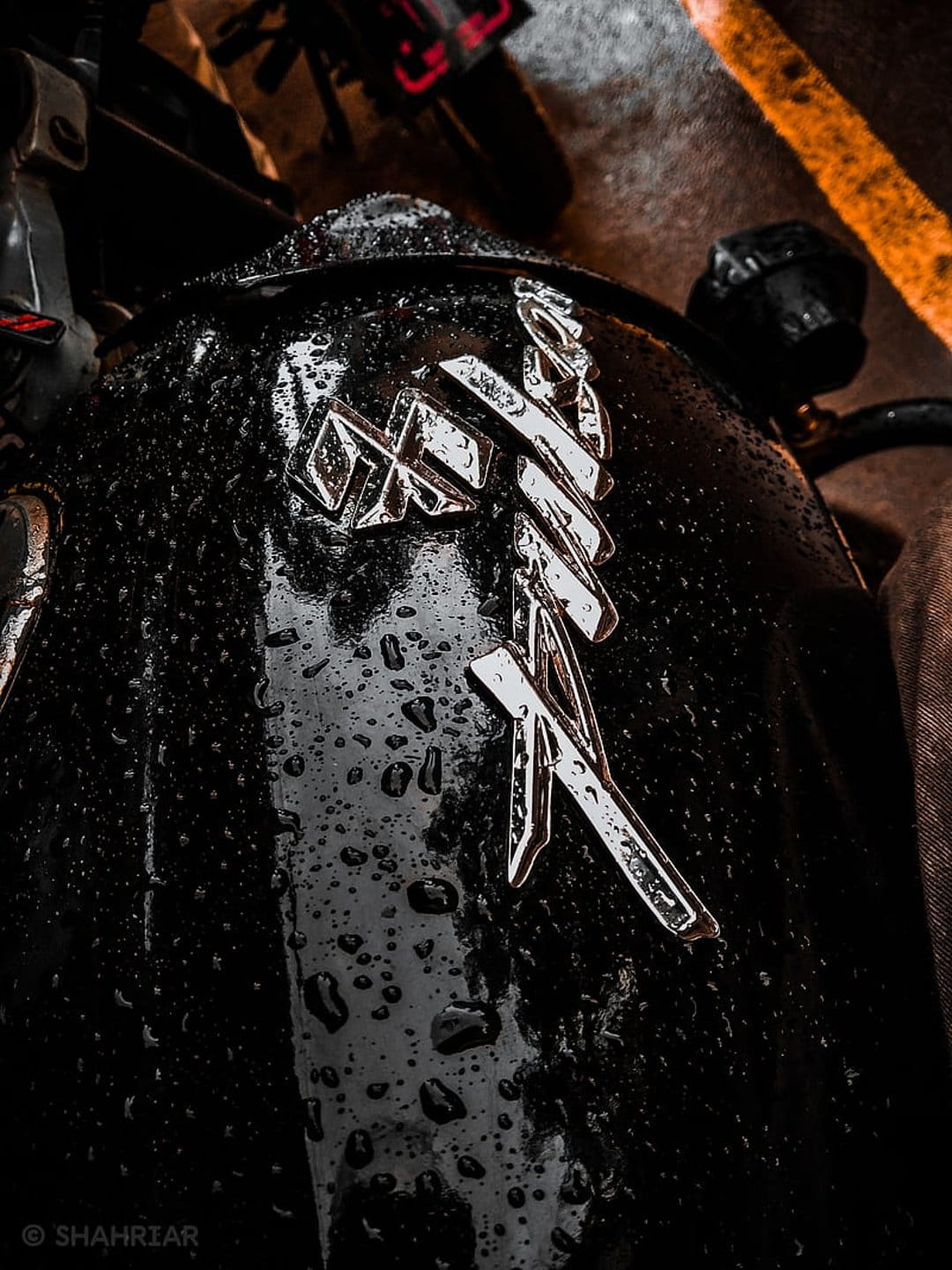 Black motorcycle fuel tank with a silver chrome emblem covered in small, clear raindrops after a rain shower.