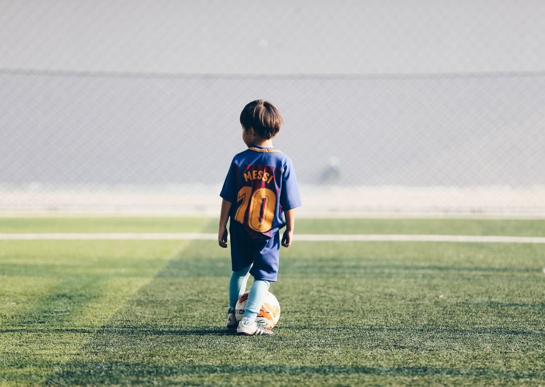 Young boy wearing a blue number ten soccer jersey stands on a green field with a football at his feet.