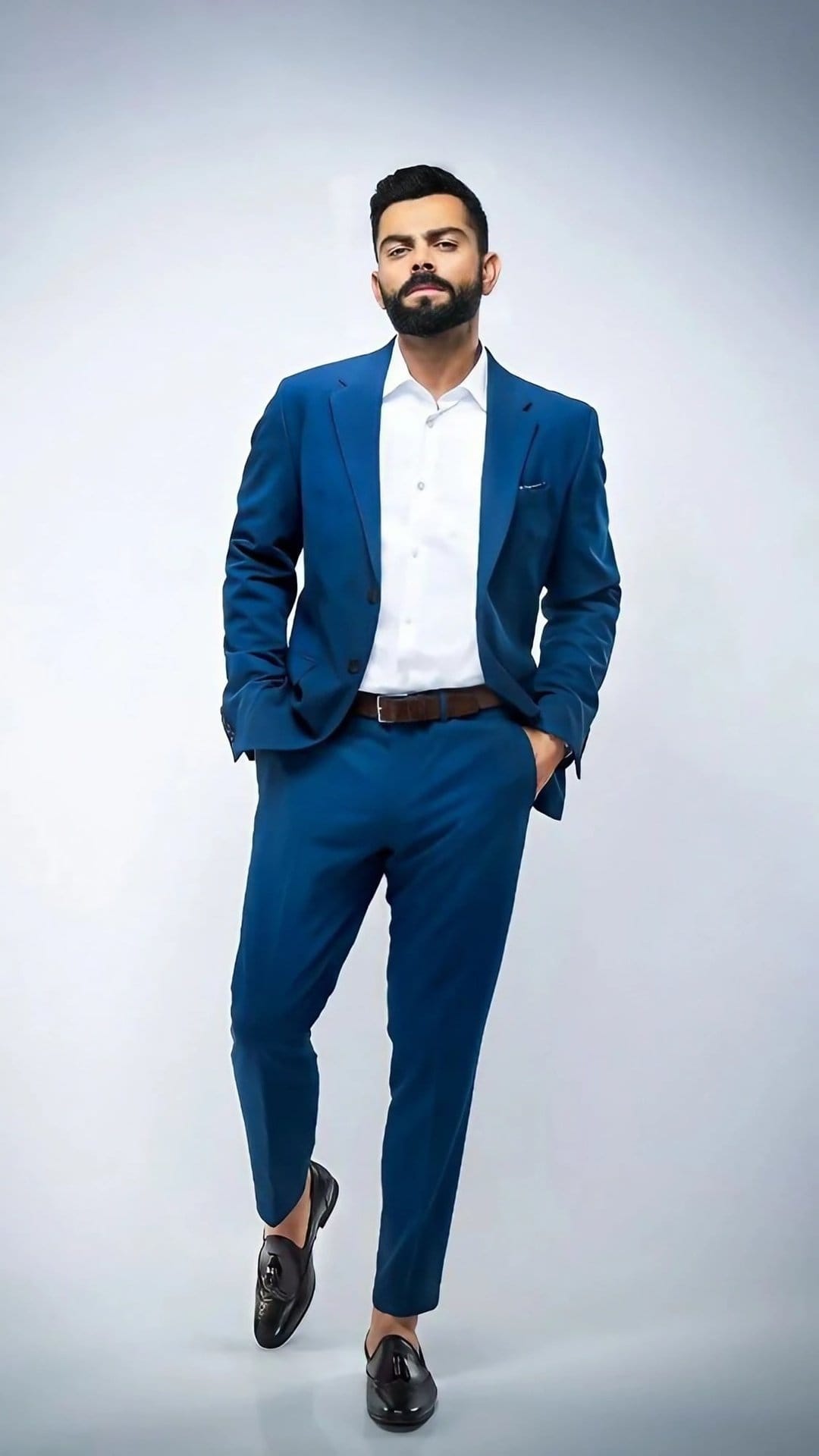 Bearded man in a tailored navy blue suit with hands in pockets stands against a clean, light studio background.