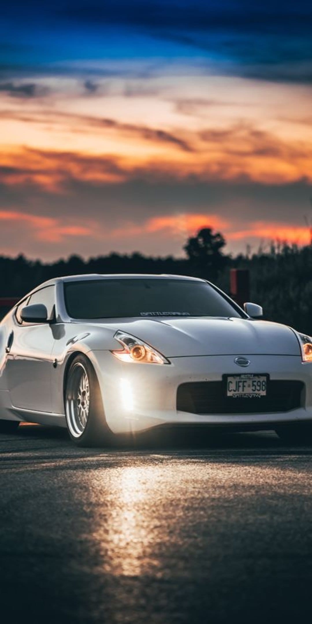 White Nissan 370Z sports car with glowing headlights on an asphalt street during a vibrant orange sunset sky.