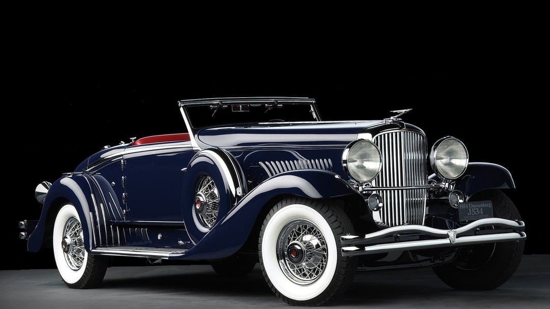 Navy blue 1934 Duesenberg Model J Roadster with a red interior and chrome details on a black background.