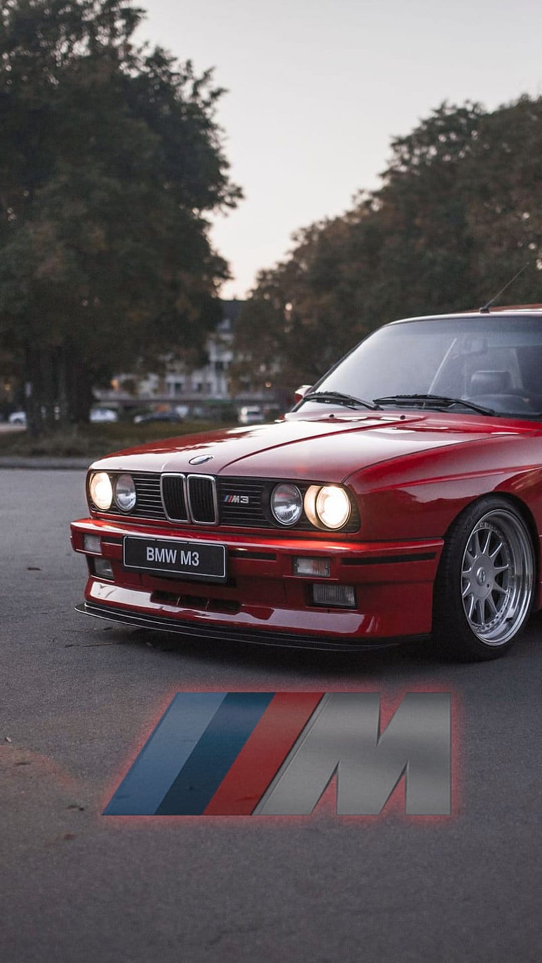 Red BMW M3 front grille and headlights featuring a vintage badge and tri-color logo on dark asphalt below.