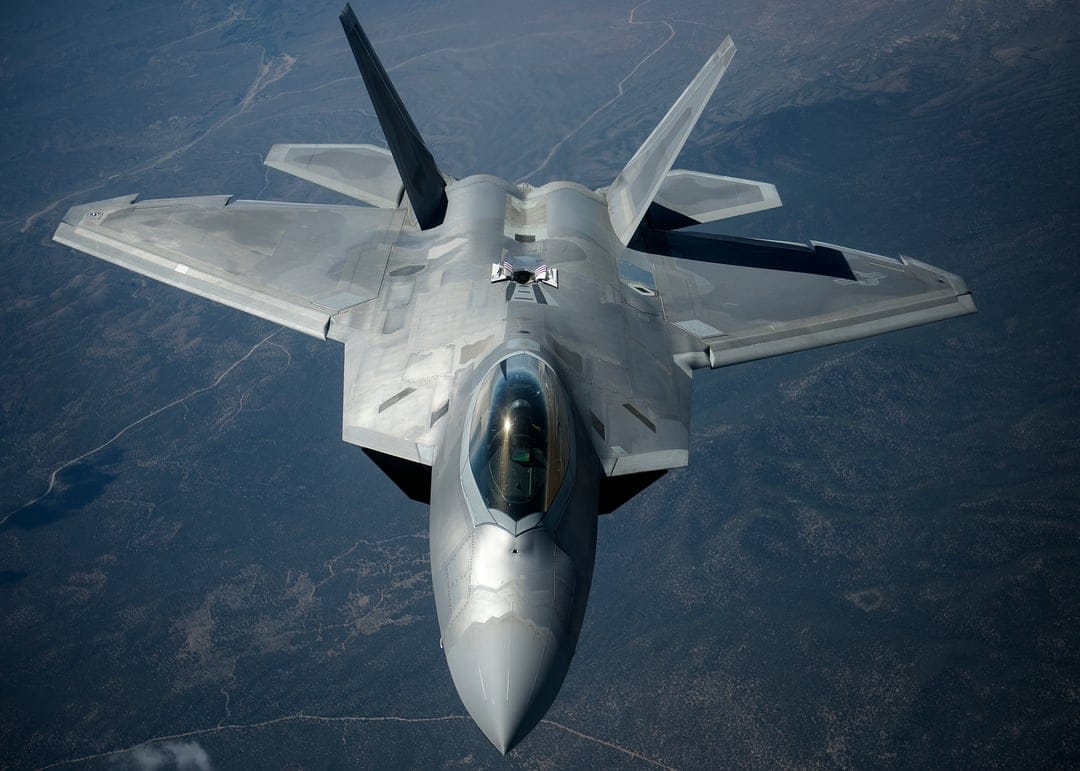 F-22 Raptor stealth fighter jet flies over a mountainous landscape as seen from a high-angle overhead perspective.