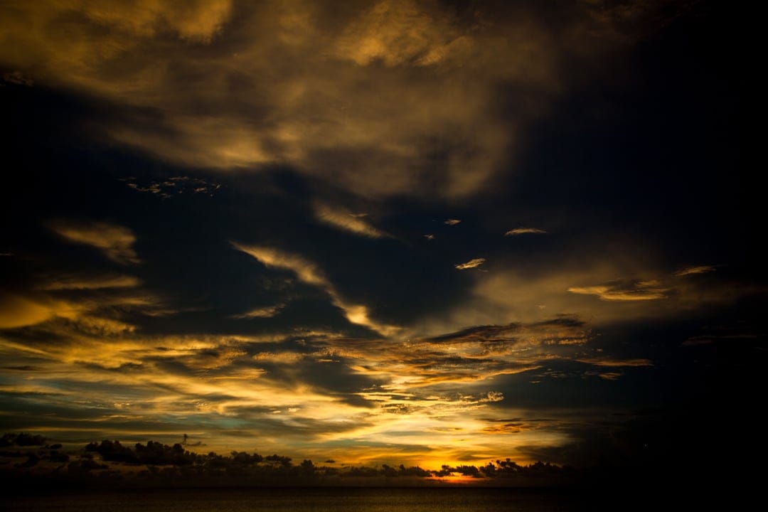 Golden clouds hover over a dark calm ocean horizon during a dramatic coastal sunset in a vertical landscape.