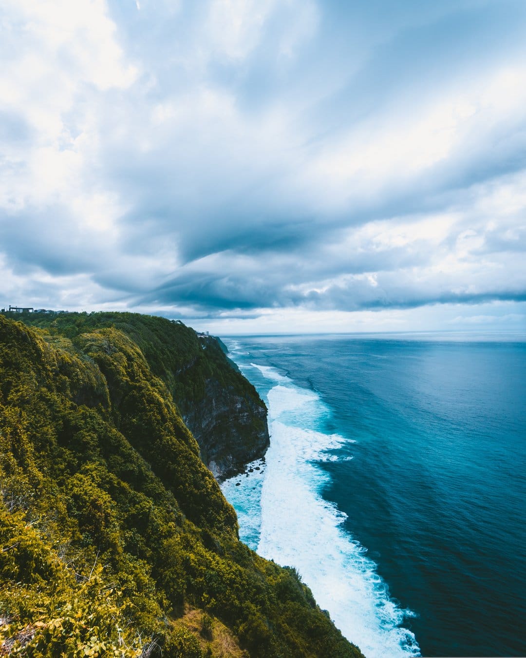Lush green cliffs overlook swirling turquoise ocean waves under a moody sky in this vertical coastal landscape.