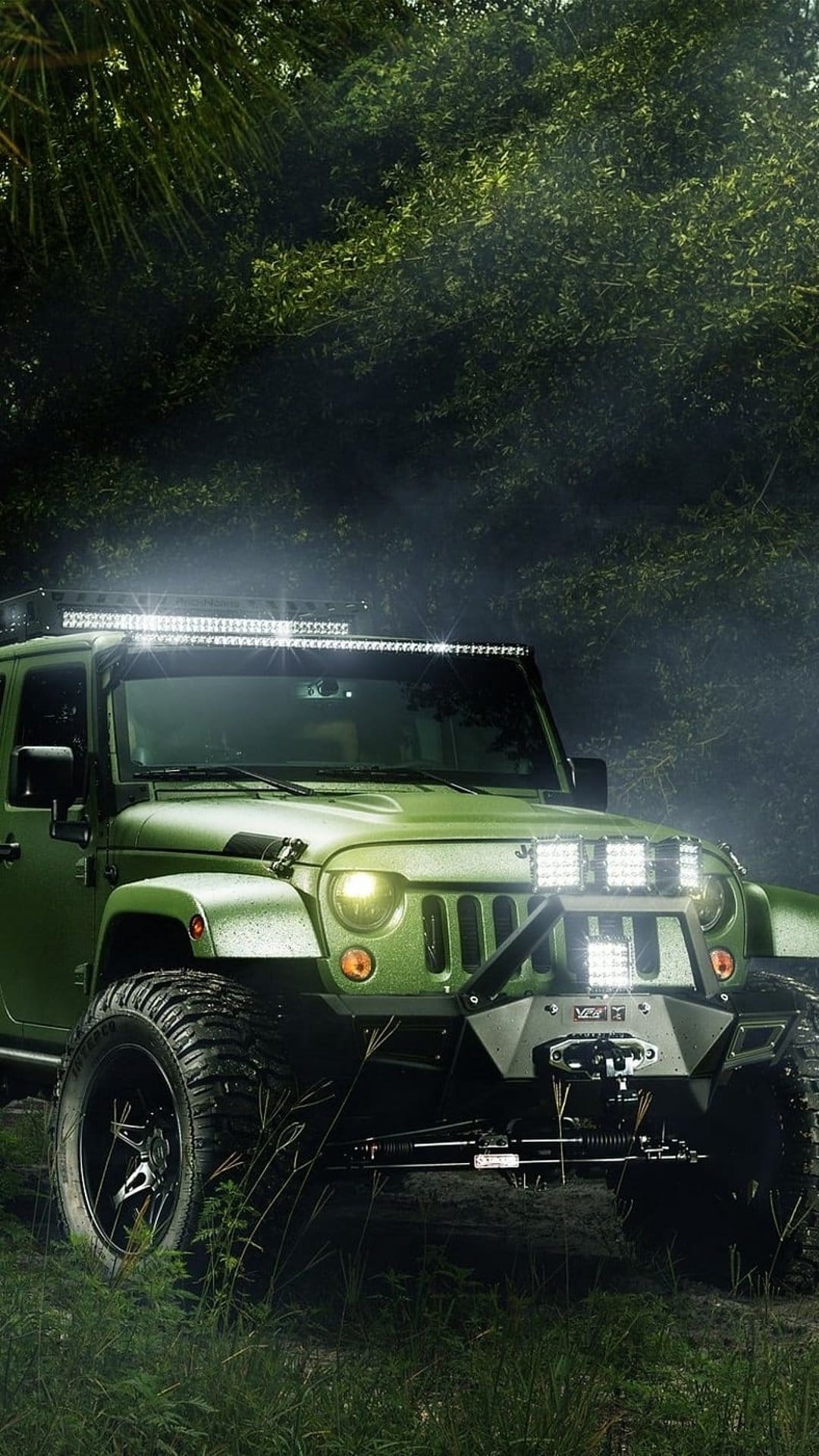 Green Jeep Wrangler with luminous LED light bars parked on a forest trail amidst thick fog and pine trees.
