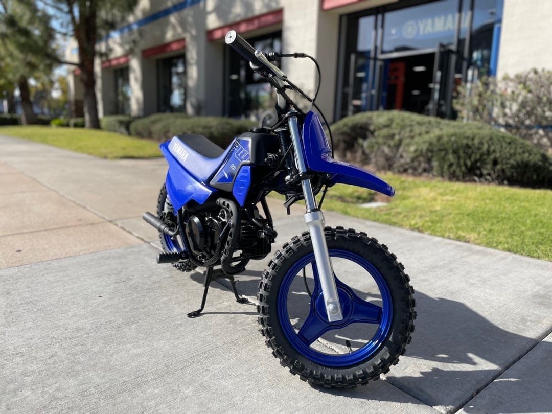 Blue Yamaha PW50 youth dirt bike with knobby tires parked on a sidewalk in front of a dealership.