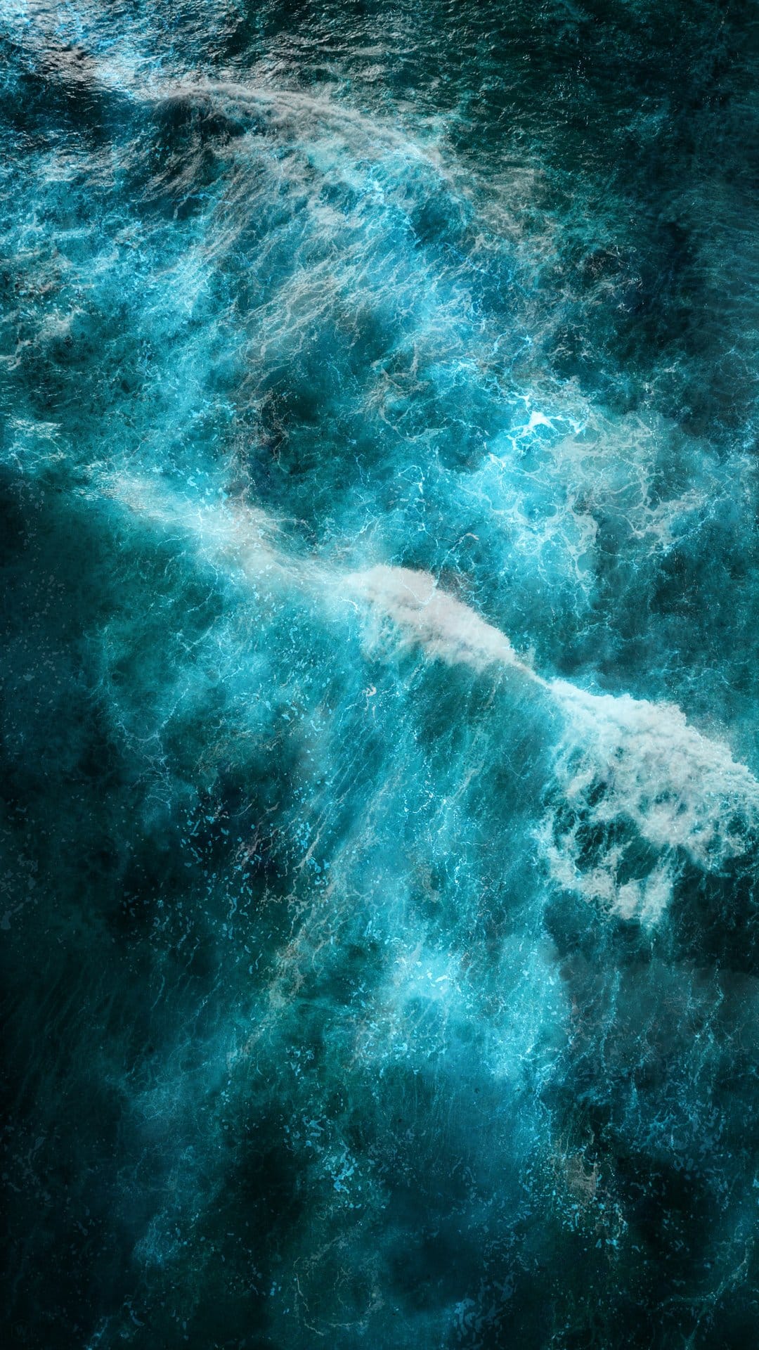Turquoise ocean waves crash together creating white sea foam and intricate water textures seen from directly above.