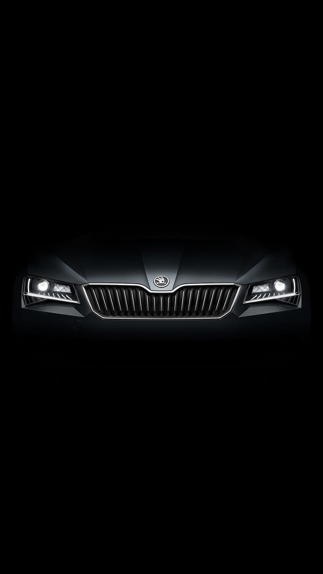 Skoda car front grille and glowing LED headlights emerge from a pitch-black background in a low-angle shot.
