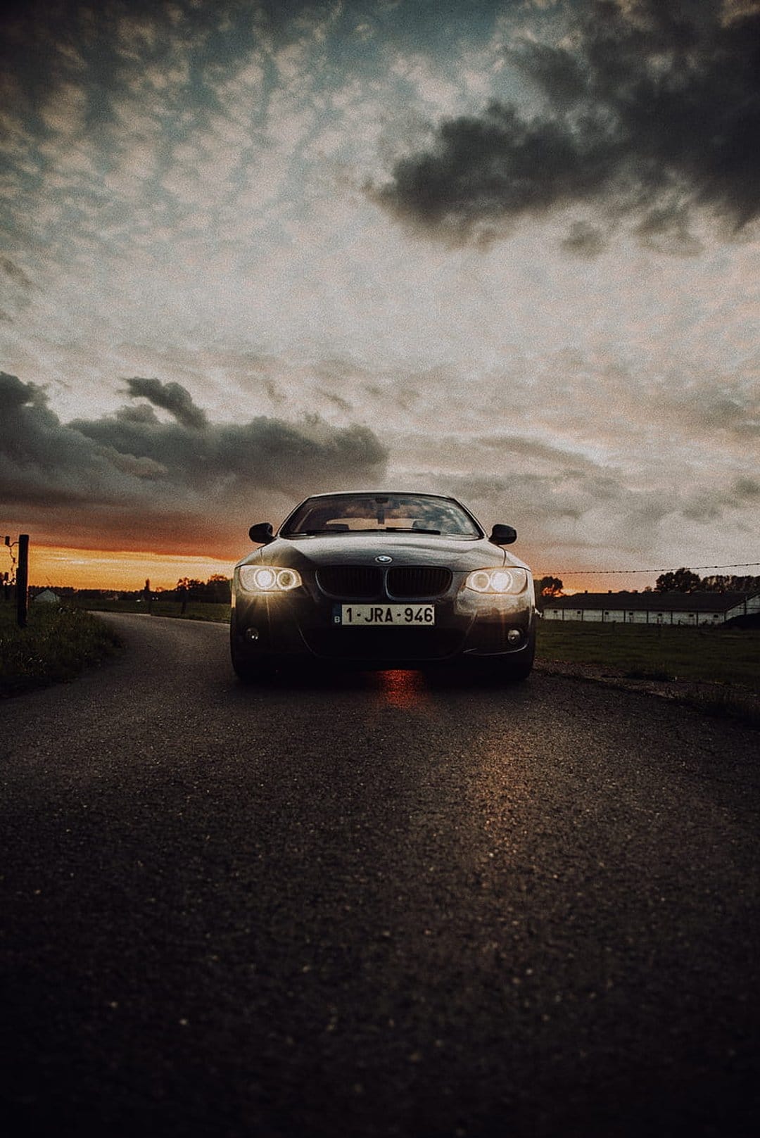 BMW car with lit headlights driving on an asphalt road at sunset under a dark and cloudy moody sky.