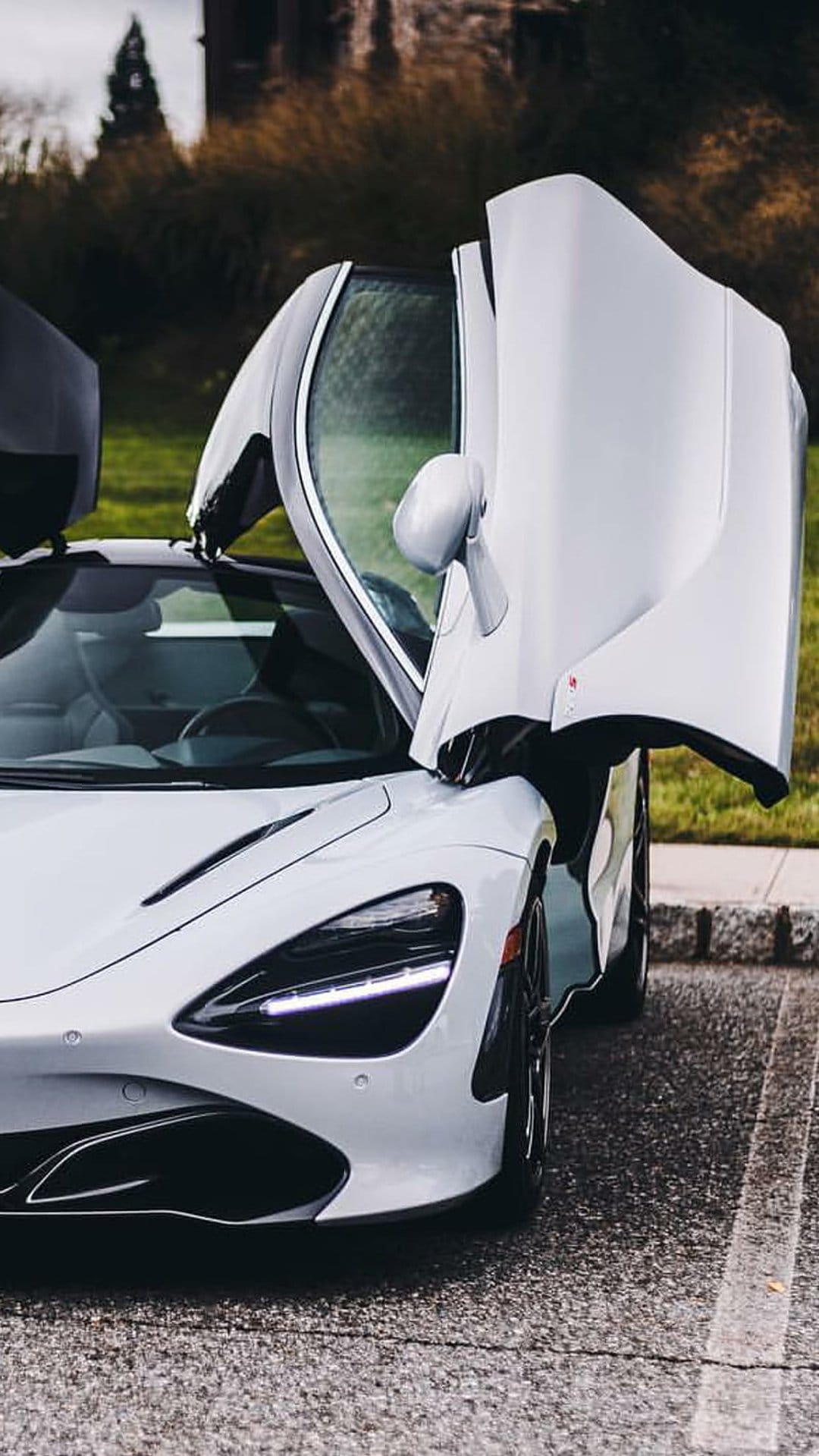 White McLaren 720S supercar with dihedral doors open parked on an outdoor road under bright natural light.