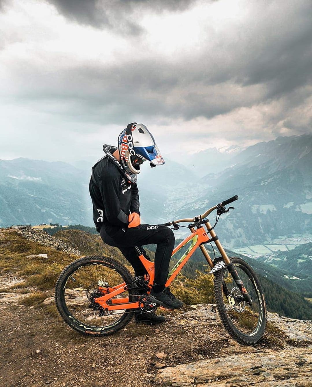 Mountain biker in full gear sits on a bright orange bike atop a rocky cliff overlooking misty peaks and grey clouds.