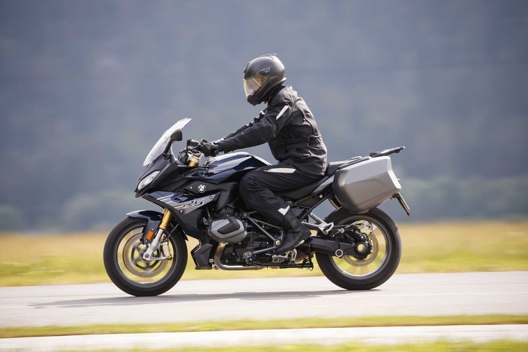 A motorcyclist in black gear riding a dark blue BMW R 1250 RS on an open road with a motion blur background.