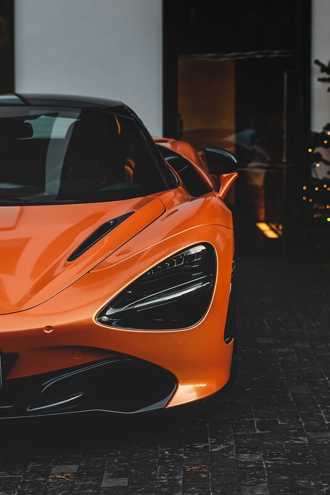 Orange McLaren 720S supercar parked on dark cobblestone with glowing holiday tree lights in the background.