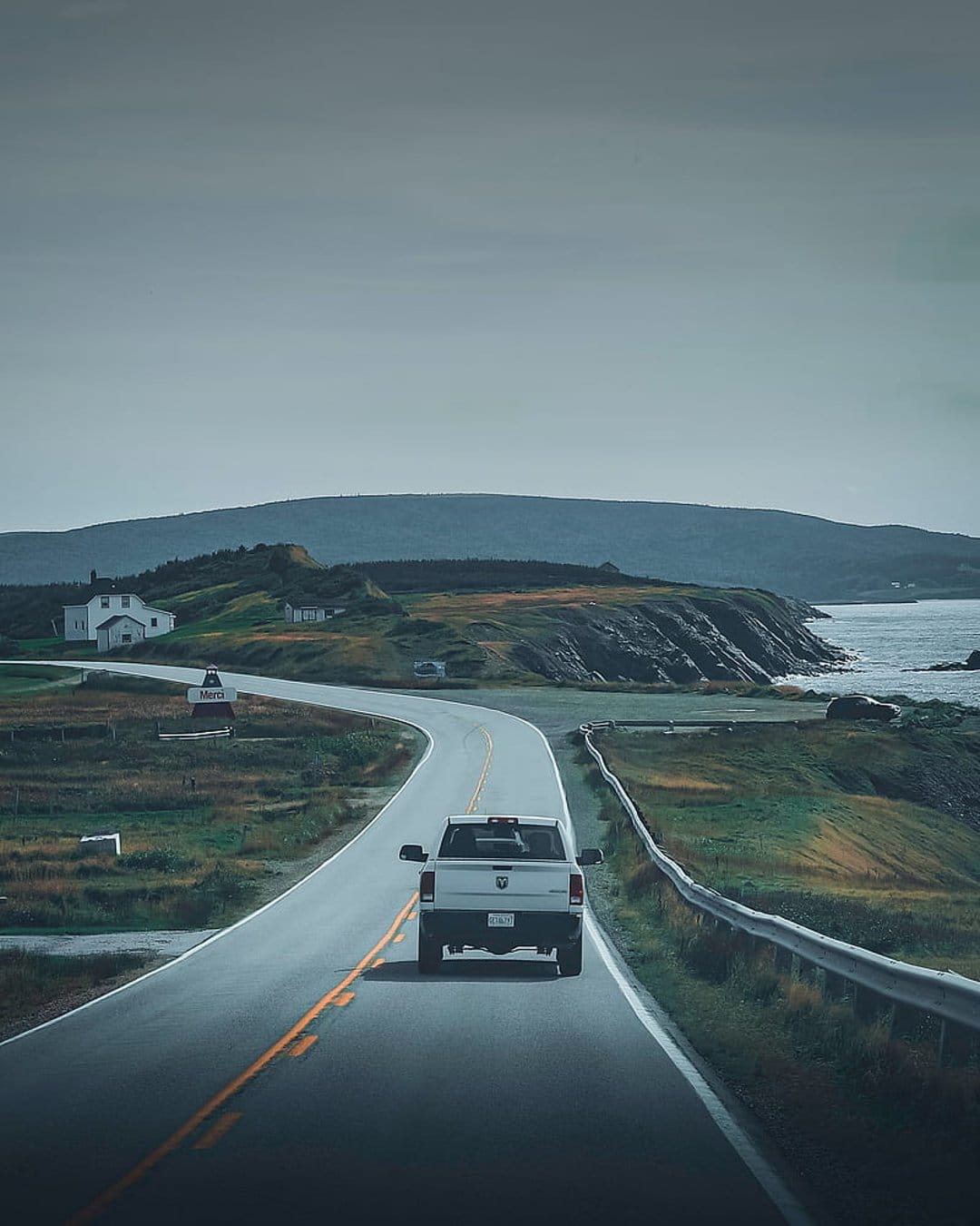 White pickup truck drives along a winding seaside highway past rocky cliffs and misty ocean views under grey skies.
