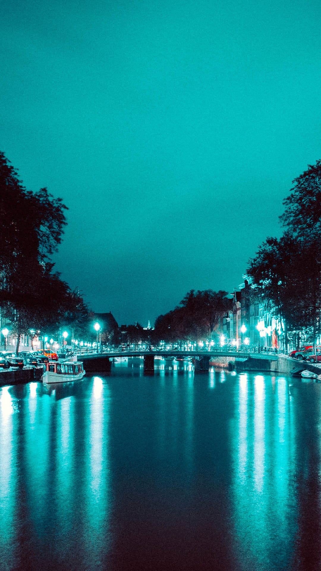 Amsterdam canal bridge illuminated by teal lights reflecting in dark water with docked boats and bare winter trees.